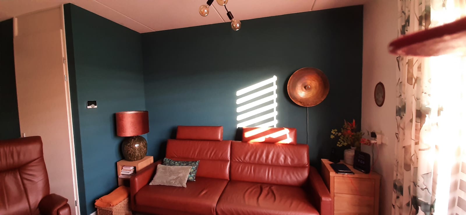 Een woonkamer met een diep petrolblauwe muur achter een roodleren bank
